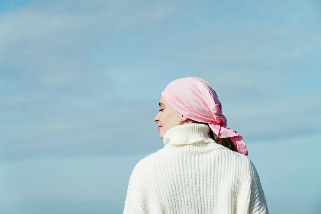 A imagem mostra uma mulher em pé ao ar livre contra um céu azul claro, vestindo um suéter branco e um lenço de cabeça rosa, representando o congelamento de óvulos para pessoas em tratamento oncológico.