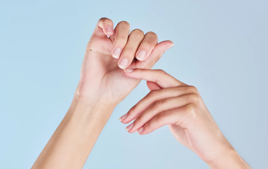 Duas mãos, uma segurando a outra, com unhas bem cuidadas e um fundo azul claro. Imagem faz alusão ao autocuidado sem culpa.