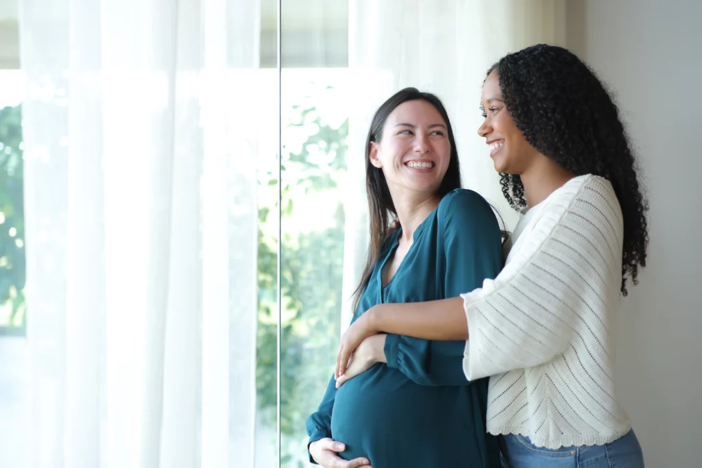 Duas mulheres sorridentes, uma delas grávida e com a mão da outra em sua barriga, em um momento que pode ter sido possibilitado pela inseminação artificial com sêmen de doador.