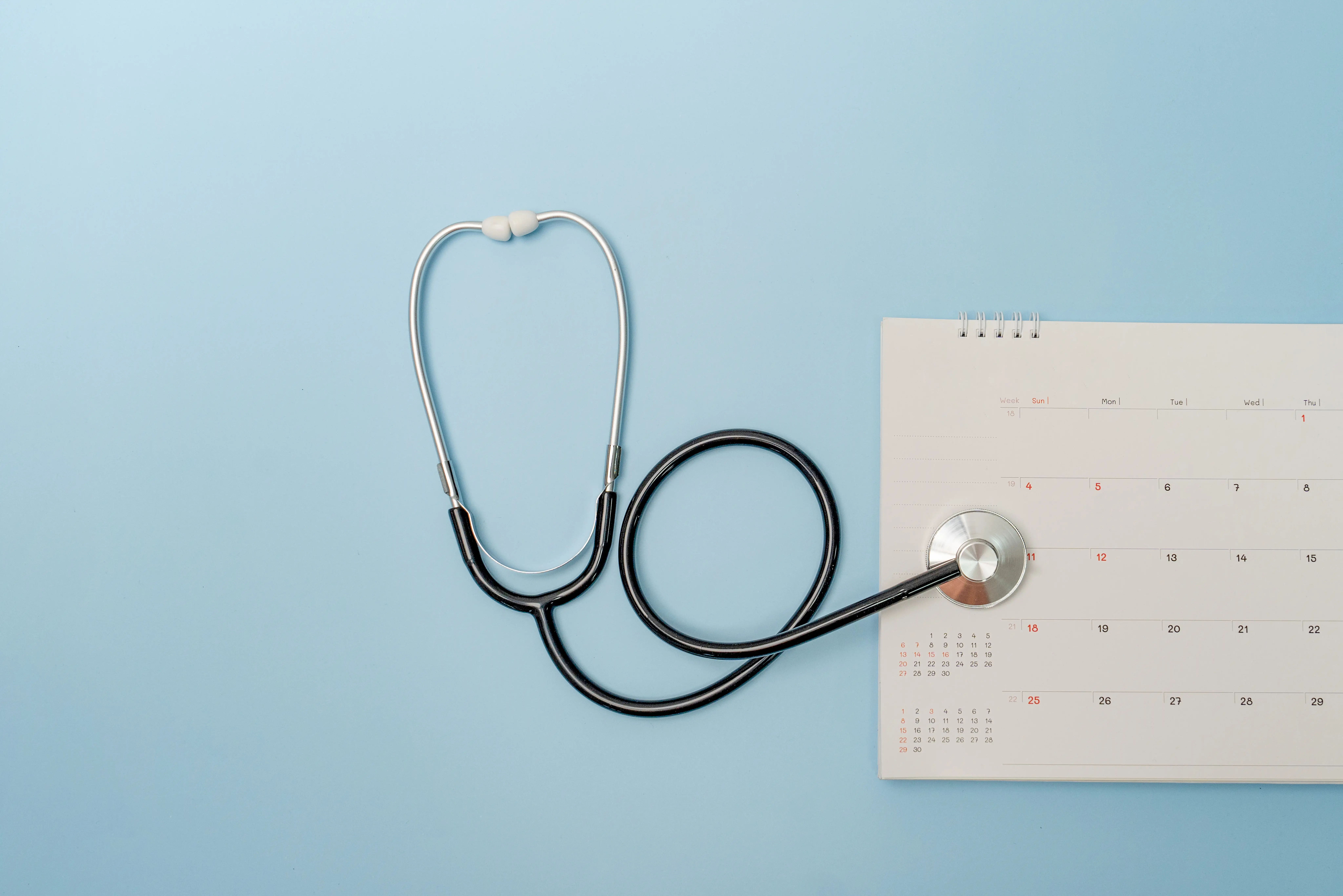 Um estetoscópio preto e prateado está ao lado de um calendário branco em um fundo azul-claro.