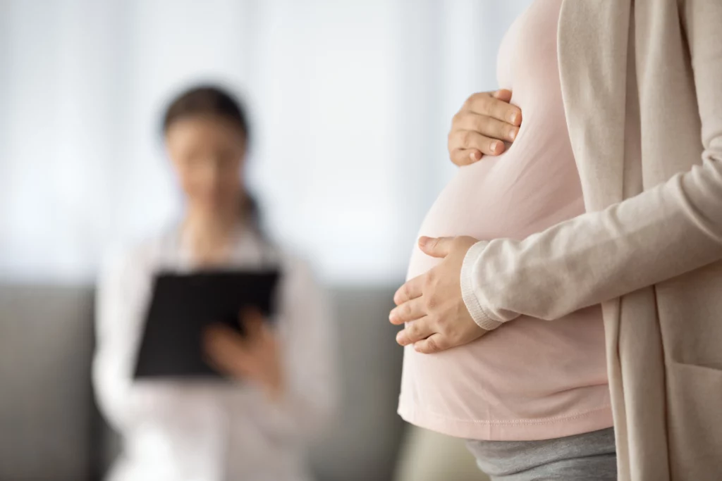 Uma mulher grávida com as mãos na barriga, com uma médica borrada ao fundo.